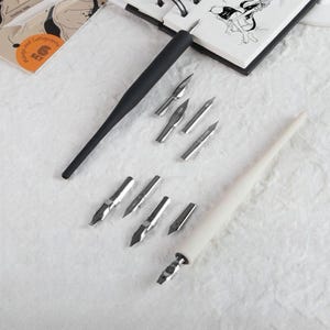 May include: A calligraphy set featuring one black pen holder, one white pen holder, and eight interchangeable metal nibs of various shapes and sizes. The set is displayed alongside a sketchbook with manga and calligraphy text visible.