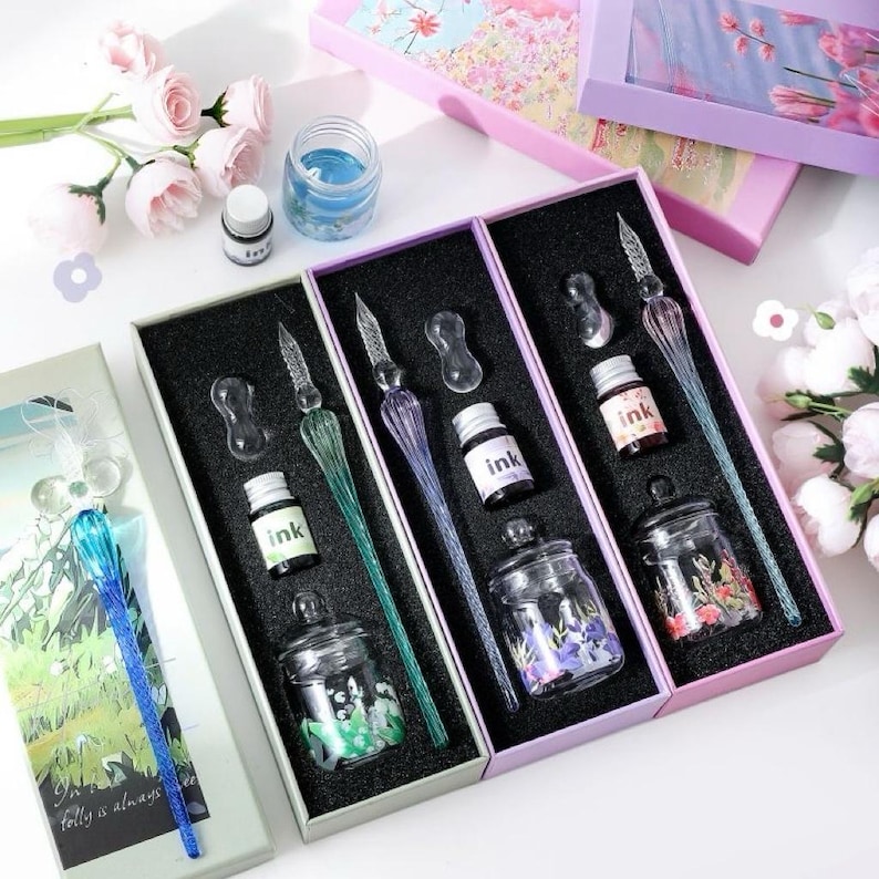 K&ouml;nnte beinhalten: Kalligraphie-Sets in Schachteln, jeweils mit Glasfeder, Tintenfass und Tintenf&auml;sschen. Die Stifte haben ein dekoratives Design. Die Tintenf&auml;sser sind mit floralen Mustern verziert. Die Schachteln sind in Grau-, Lila- und Rosat&ouml;nen gehalten.