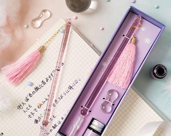 May include: A glass dip pen set featuring two transparent pink glass pens with twisted nibs and light pink tassels. The set includes a small bottle of ink with a purple label and a clear glass pen rest, all housed in a rectangular purple box.