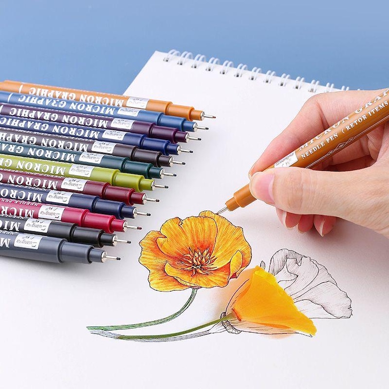 May include: A collection of Micron Graphic needle pens in various colors, with one pen in use drawing a detailed illustration of a flower. The pens are arranged on a white spiral-bound notepad, showcasing their fine-tip capabilities.