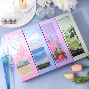 K&ouml;nnte beinhalten: Vier dekorative Schachteln mit Landschafts- und Blumendesigns. Die Schachteln sind in Rosa-, Blau- und Gr&uuml;nt&ouml;nen gehalten, mit dem Wort "Heart" auf jeder Schachtel. Die Schachteln sind auf einer hellblauen Oberfl&auml;che angeordnet, mit Glasstiften und kleinen Gl&auml;sern.