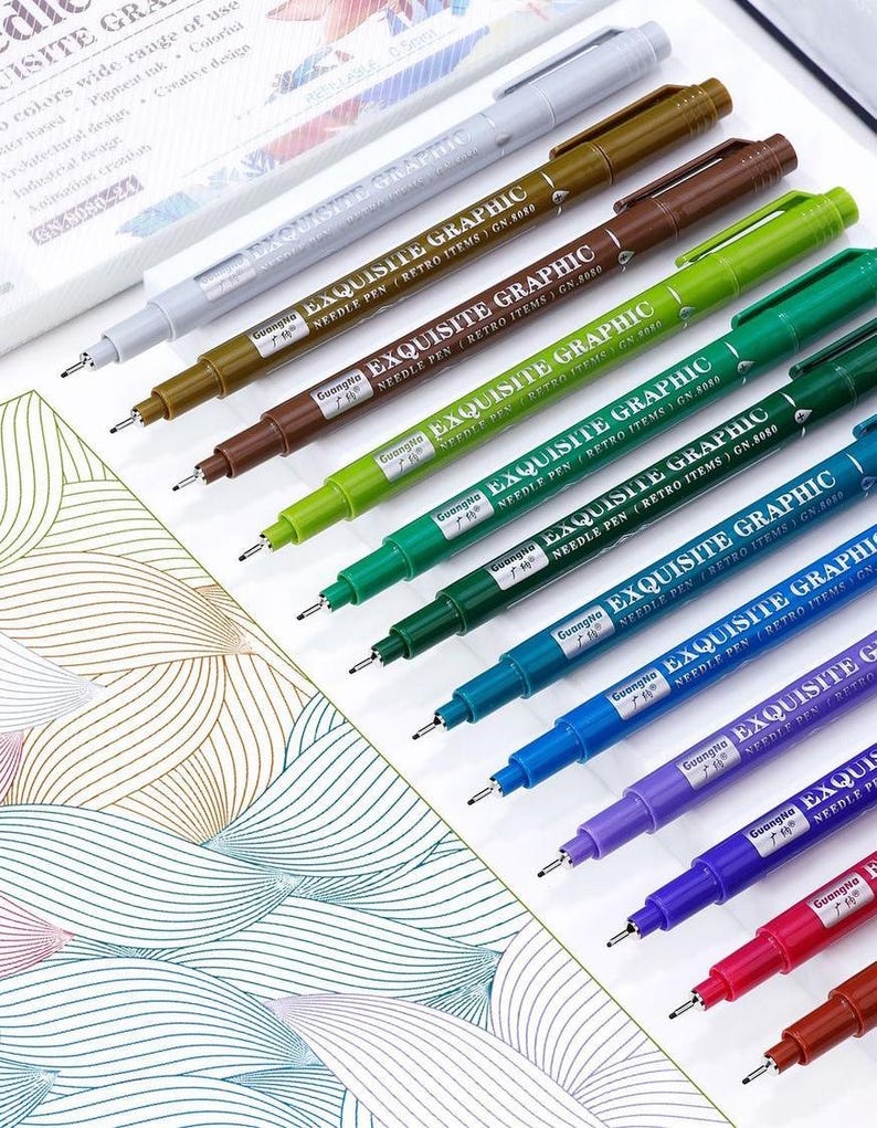May include: A collection of colorful Exquisite Graphic needle pens. The pens are arranged diagonally, showcasing a range of colors including brown, green, blue, purple, and red. Each pen has a fine tip and the text "EXQUISITE GRAPHIC" printed on the barrel.