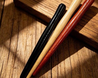 May include: Three cylindrical wooden sticks arranged diagonally. One stick is finished in a glossy black, one in a natural light tan wood tone, and one in a deep reddish-brown wood grain. Each stick features a smooth, tapered, rounded end.