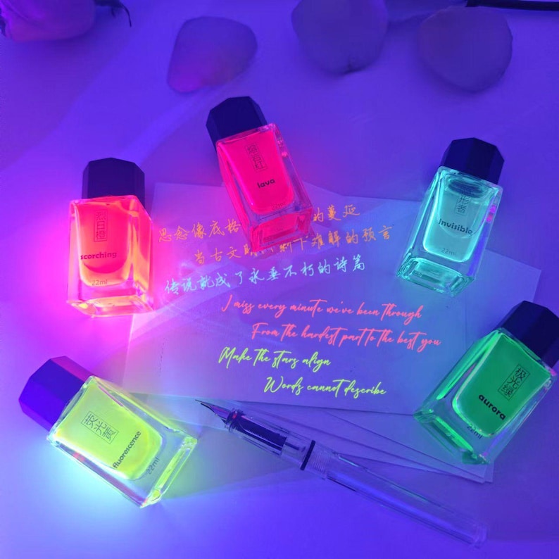 May include: Five small, rectangular perfume bottles with black caps and glowing neon colors: orange, pink, green, and yellow. The bottles are labeled with words like "scorching" and "aurora". A clear pen and a handwritten note are also visible.