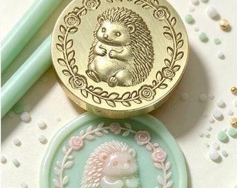May include: A circular brass wax seal stamp featuring an engraved hedgehog surrounded by a floral wreath. Below it, a light green wax seal impression displays the same hedgehog and floral design in raised, pale pink and green relief.