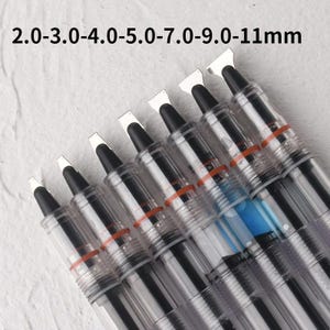 May include: A set of seven calligraphy pens with clear plastic barrels and black internal components. Each pen features a flat metal nib with varying widths labeled 2.0, 3.0, 4.0, 5.0, 7.0, 9.0, and 11mm. The pens are arranged diagonally.
