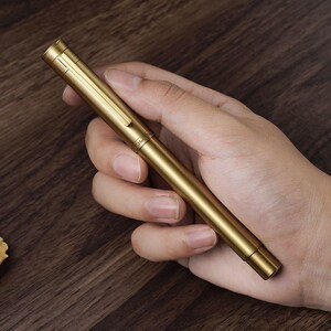 Puede incluir: Un bol&iacute;grafo de color dorado con un clip y un cuerpo cil&iacute;ndrico. El bol&iacute;grafo se sostiene en una mano y el fondo es una superficie de madera oscura. El bol&iacute;grafo parece ser de metal y tiene un acabado mate.