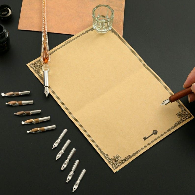 Op de afbeelding: Een kalligrafieset met een transparante glazen dompelpen met een gedraaide houder, een kleine glazen inktpot en tien verwisselbare metalen penpunten. Een vel beige briefpapier met een decoratieve zwarte rand en een sleutelpictogram is inbegrepen.