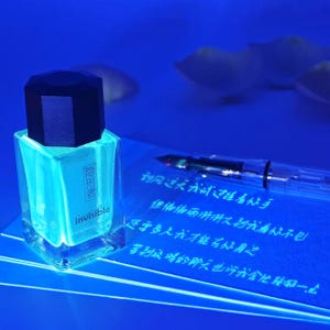 May include: A square glass bottle of glowing turquoise ink with a dark blue cap. The bottle has the word "invisible" and Chinese characters on the front. A clear fountain pen and handwritten text are in the background.