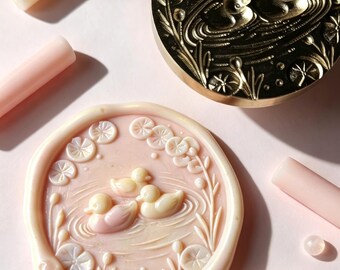 May include: A round brass wax seal stamp featuring an engraved design of two ducks swimming in a pond with lily pads. Beside it is a matching pale pink wax seal impression showing the same duck and lily pad motif, surrounded by several cylindrical wax sticks and small round wax beads.