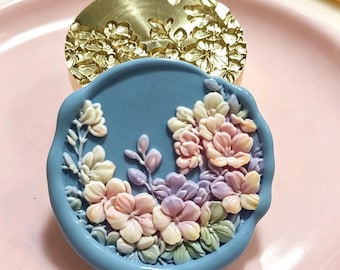 May include: A circular blue wax seal featuring a raised, multi-colored floral relief design in shades of pink, purple, and cream, positioned in front of a gold-toned metal stamp head with an engraved floral pattern on its surface.
