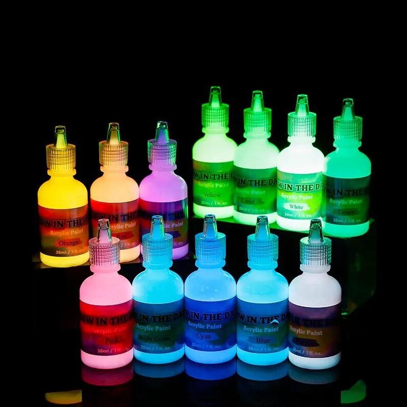 Puede incluir: Una colecci&oacute;n de doce botellas de pl&aacute;stico peque&ntilde;as con pintura acr&iacute;lica que brilla en la oscuridad, cada una etiquetada con 30 ml. Las botellas est&aacute;n dispuestas en dos filas sobre un fondo negro, mostrando varios colores vibrantes como amarillo, naranja, rosa, verde, azul y blanco.