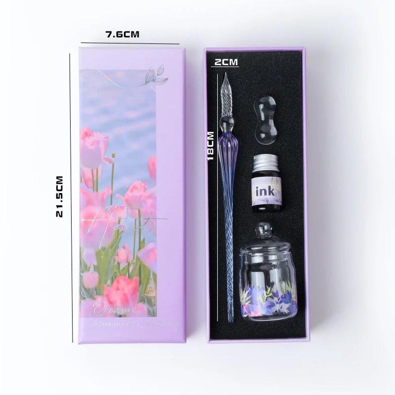 K&ouml;nnte beinhalten: Ein lila Kalligraphie-Set mit einem Glas-Dip-Pen (18 cm), einer Tintenflasche, einem Tintenfass und einem kleinen Glas. Die Box ist 21,5 cm lang und 7,6 cm breit und hat ein Blumendesign. Die Tintenflasche ist mit "ink" beschriftet.