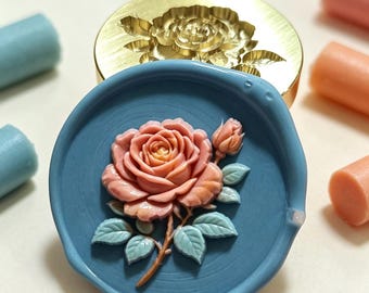 May include: A circular blue wax seal featuring a raised, multi-colored rose design with pink petals and light green leaves. Behind the seal sits a round brass stamp head engraved with a matching rose pattern, surrounded by several small wax sticks.