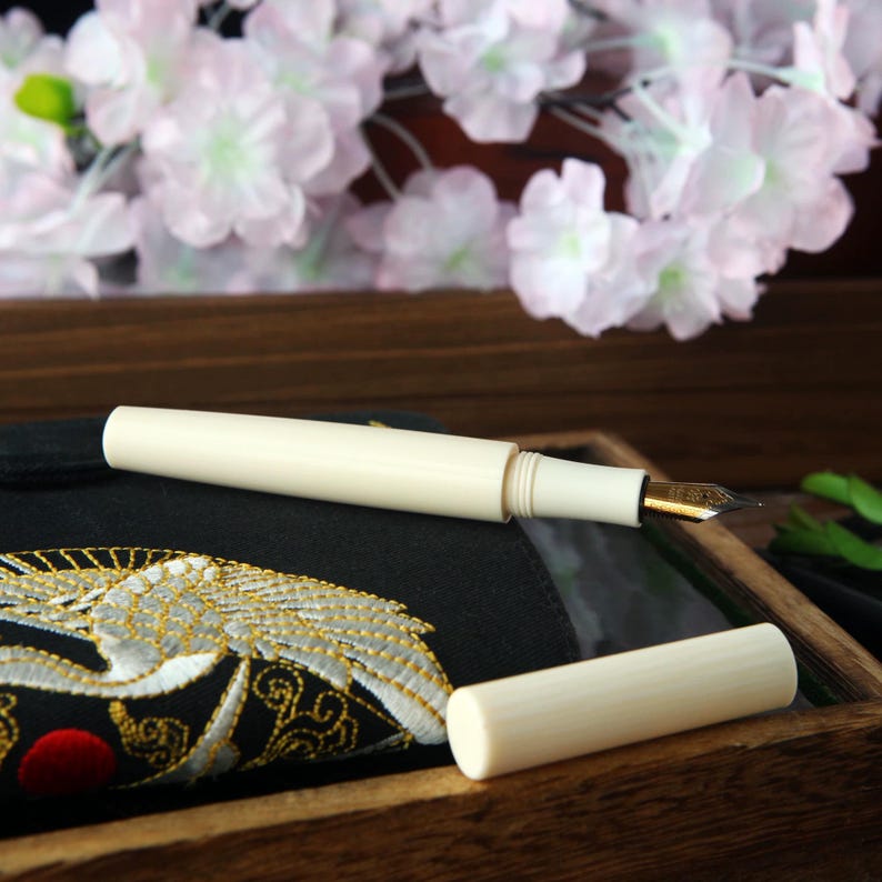May include: A white fountain pen with a gold nib, closed and ready to write. The pen is on a black fabric with a gold embroidered crane design.