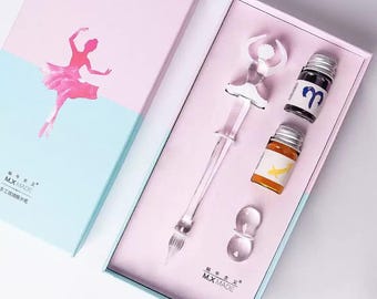 May include: A glass dip pen with a ballerina figure handle, a clear glass pen rest, and two small glass bottles of ink, one with a blue Aries symbol and one with a yellow fish graphic, all housed in a pink and light blue gift box labeled The Ballet Girl.