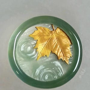 May include: A round, jade-green pendant with a gold-colored maple leaf design. The leaf is centered on a light green background with a water ripple effect. The pendant has a polished, glossy finish.