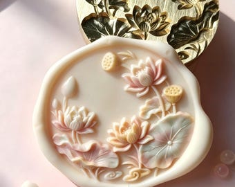 May include: A circular brass wax seal stamp featuring an intricate lotus flower relief design, paired with a matching cream-colored wax seal impression showing pink-tinted lotus blossoms, buds, and leaves, surrounded by several pale wax sticks.