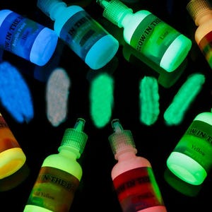 Puede incluir: M&uacute;ltiples frascos de 30 ml de pintura acr&iacute;lica que brilla en la oscuridad dispuestos sobre una superficie negra. Cada frasco tiene una etiqueta con el nombre del color y muestras de pintura debajo, mostrando pigmentos luminosos azules, blancos, verdes y amarillos.