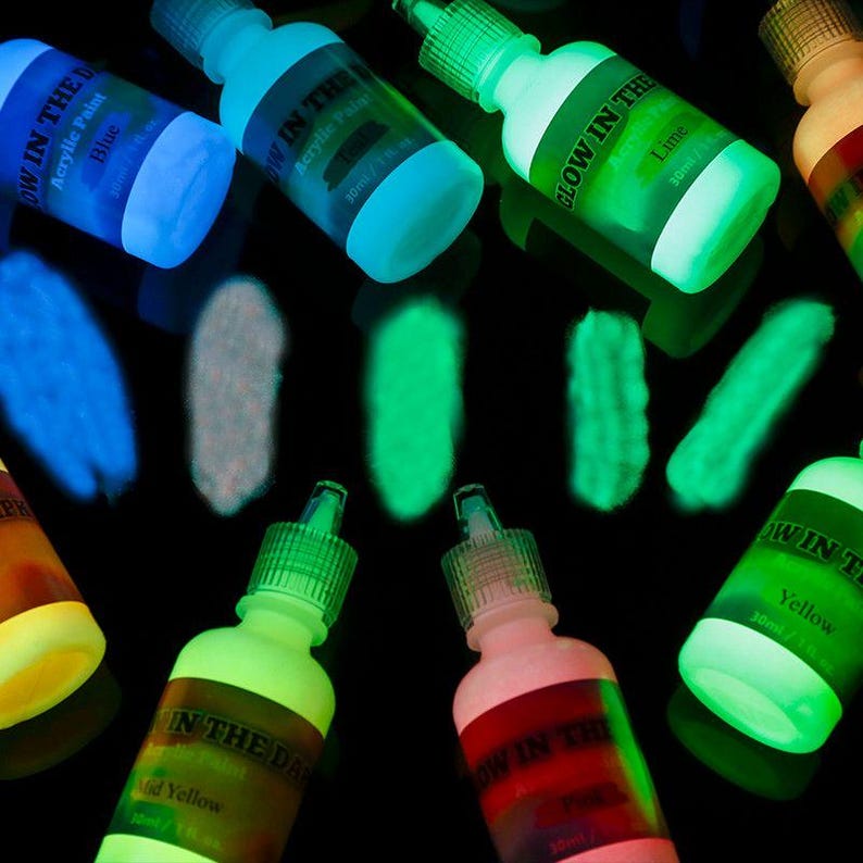 Puede incluir: M&uacute;ltiples frascos de 30 ml de pintura acr&iacute;lica que brilla en la oscuridad dispuestos sobre una superficie negra. Cada frasco tiene una etiqueta con el nombre del color y muestras de pintura debajo, mostrando pigmentos luminosos azules, blancos, verdes y amarillos.