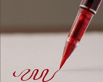 May include: A red brush pen with a transparent barrel and cap, featuring a flexible felt tip that is currently drawing a continuous, wavy red line on a light-colored surface. The ink inside the pen barrel is visible through the clear plastic casing.
