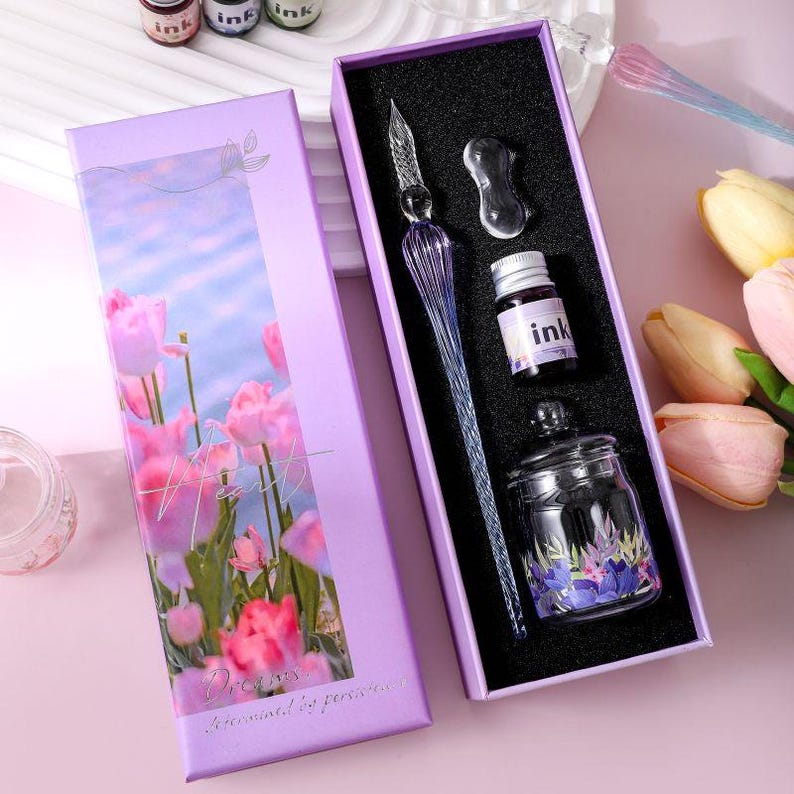 K&ouml;nnte beinhalten: Ein Kalligraphie-Set in einer lila Schachtel mit Blumendesign. Das Set enth&auml;lt einen Glasstift, eine Tintenflasche, ein Tintenfass und eine Stifthalterung. Die Schachtel zeigt Tulpen mit dem Wort "Heart" und dem Satz "Dreams determined by passion".