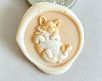 May include: A circular wax seal featuring a raised, detailed relief of a sleeping corgi dog with its paws tucked in. The seal is light beige with a smooth, glossy finish. A metal stamp with the same corgi design is positioned directly above the seal.