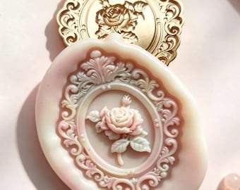 May include: A pale pink wax seal featuring a central rose relief inside an ornate oval frame, positioned partially over a gold-colored metal stamp with a matching rose design and intricate filigree border, creating a layered decorative effect.