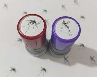 May include: Two circular self-inking stamps, one with a red casing and one with a purple casing, each featuring a white top label displaying a detailed black line drawing of a mosquito, positioned against a white background with mosquito patterns.