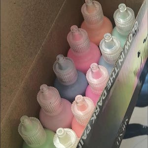 Puede incluir: Una caja de cart&oacute;n que contiene varios frascos de pl&aacute;stico peque&ntilde;os con pintura que brilla en la oscuridad. Los frascos tienen tapas puntiagudas transparentes y vienen en varios colores pastel, incluyendo rosa, azul, verde y melocot&oacute;n. La caja tiene la etiqueta GLOW IN THE DARK PAINT.