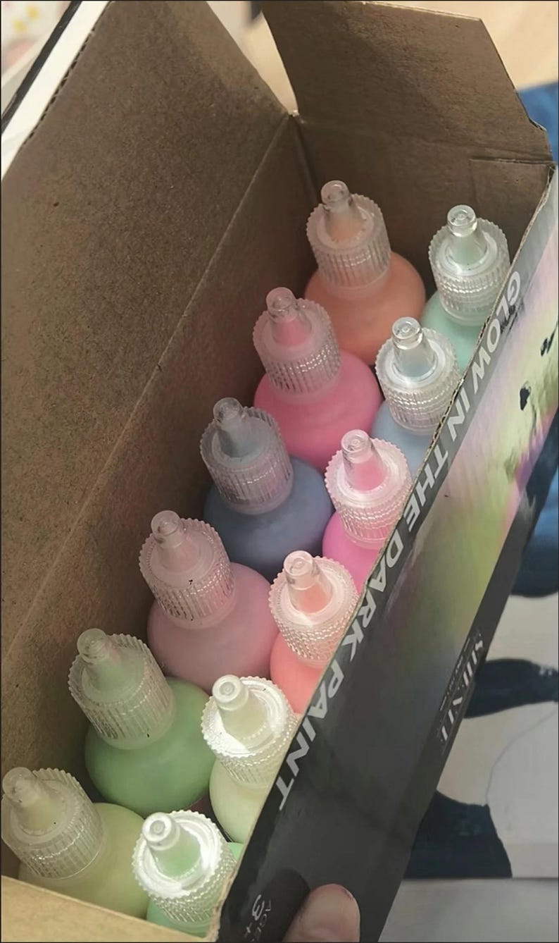 Puede incluir: Una caja de cart&oacute;n que contiene varios frascos de pl&aacute;stico peque&ntilde;os con pintura que brilla en la oscuridad. Los frascos tienen tapas puntiagudas transparentes y vienen en varios colores pastel, incluyendo rosa, azul, verde y melocot&oacute;n. La caja tiene la etiqueta GLOW IN THE DARK PAINT.