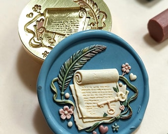 May include: A circular brass wax seal stamp featuring an engraved design of a scroll, a feather quill, and a key, paired with a blue wax seal impression showing the same detailed motif in relief, accented with small pink flowers and hearts.
