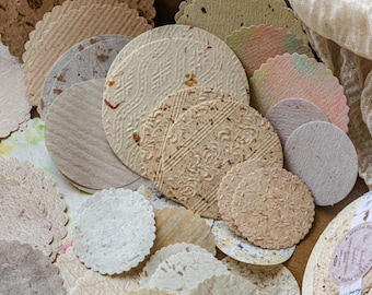 May include: A collection of circular paper coasters with scalloped edges, featuring various textures, neutral tones, and embedded botanical elements. Some pieces display embossed patterns, while others show subtle watercolor washes and flecked fibers.