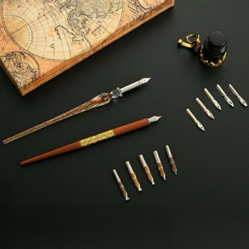 Op de afbeelding: Een kalligrafieset bestaande uit een glazen kroontjespen, een bruine houten pen met een goudkleurige metalen ring, een kleine zwarte inktpot met een goudkleurige dop en tien verwisselbare metalen penpunten in diverse vormen.