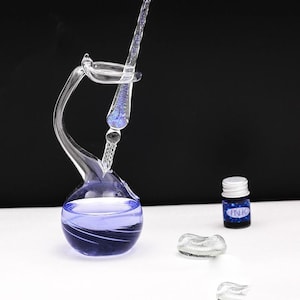 Könnte beinhalten: Ein Tauchfederhalter aus klarem Glas mit einem blauen Glitzerkern ruht in einem schwanenförmigen Glastintenfaß, das mit violetter Flüssigkeit gefüllt ist. Eine kleine Flasche mit der Aufschrift INK, weißem Deckel und zwei kleine dekorative Glasteile stehen daneben.