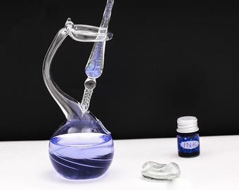 May include: A clear glass dip pen with a blue glitter core rests in a swan-shaped glass inkwell filled with purple liquid. A small bottle labeled INK with a white cap and two small clear glass decorative pieces are positioned nearby.