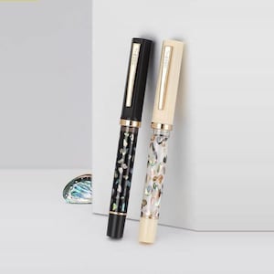 Puede incluir: Dos plumas estilogr&aacute;ficas. Una es negra con un patr&oacute;n decorativo y detalles dorados, la otra es de color crema con un patr&oacute;n similar. Ambas plumas tienen clips dorados y el texto "T-ZERO" en el clip.