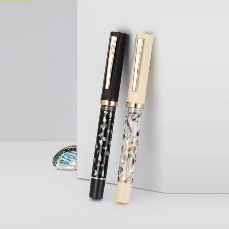 Puede incluir: Dos plumas estilogr&aacute;ficas. Una es negra con un patr&oacute;n decorativo y detalles dorados, la otra es de color crema con un patr&oacute;n similar. Ambas plumas tienen clips dorados y el texto "T-ZERO" en el clip.