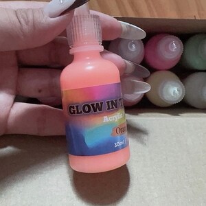 Puede incluir: Un frasco de 30 ml de pintura acr&iacute;lica naranja que brilla en la oscuridad con una tapa de boquilla de pl&aacute;stico transparente. La etiqueta tiene un fondo degradado de arco&iacute;ris con el texto GLOW IN THE DARK, Acrylic, Orange y 30ml en blanco y negro.