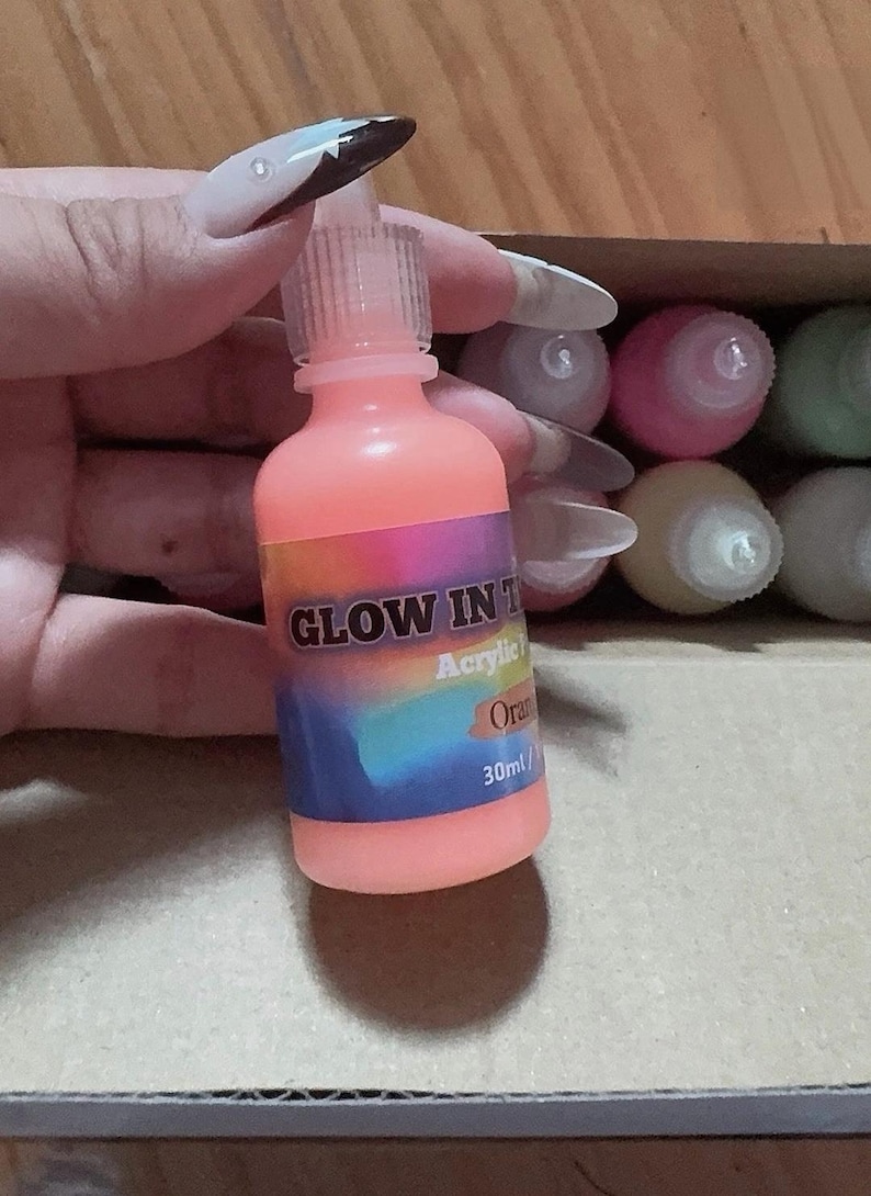 Puede incluir: Un frasco de 30 ml de pintura acr&iacute;lica naranja que brilla en la oscuridad con una tapa de boquilla de pl&aacute;stico transparente. La etiqueta tiene un fondo degradado de arco&iacute;ris con el texto GLOW IN THE DARK, Acrylic, Orange y 30ml en blanco y negro.