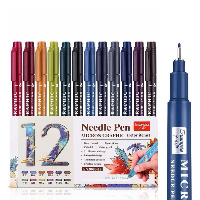 May include: A set of 12 colorful micron graphic needle pens with the text "RAPHIC" on each pen. The pens are in various colors, including red, orange, green, and blue. The packaging includes the text "Needle Pen MICRON GRAPHIC".