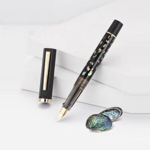 Puede incluir: Una pluma estilogr&aacute;fica negra con plum&iacute;n dorado y detalles iridiscentes. La tapa de la pluma est&aacute; quitada, revelando una banda dorada. Un par de conchas iridiscentes est&aacute;n junto a la pluma.