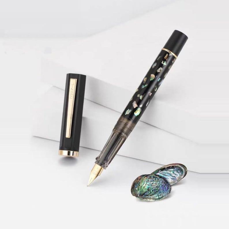 Puede incluir: Una pluma estilogr&aacute;fica negra con plum&iacute;n dorado y detalles iridiscentes. La tapa de la pluma est&aacute; quitada, revelando una banda dorada. Un par de conchas iridiscentes est&aacute;n junto a la pluma.