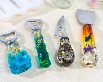 May include: A set of four kitchen tools with resin handles. Two bottle openers feature blue and green resin with gold flakes. A cheese knife has a black resin handle with embedded metal gears. A spreader features an orange and yellow resin handle with a dolphin, starfish, and seashell.