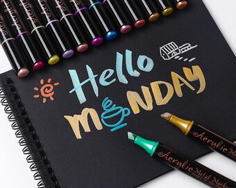 May include: A set of metallic paint markers with a variety of colors, including gold, silver, copper, and blue. The markers are arranged in a row on a black surface with the words "Hello Monday" written in metallic paint.