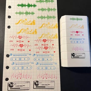 May include: A sheet of grid paper featuring various music-themed stickers including green sound waves, yellow musical notes, red heartbeats, blue playback controls, and black loading bars, alongside a stack of six white rectangular stamp blocks.