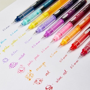 May include: A set of nine Moka gel pens with 0.5 mm tips, featuring barrels in blue, turquoise, yellow, lilac, purple, pink, orange, red, and wine red. Each pen is displayed next to its corresponding ink color swatch and a small planet doodle.