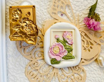 May include: A rectangular gold-toned metal wax seal stamp featuring a detailed rose design, paired with a white wax seal impression showing a pink and green rose motif, resting on a beige paper butterfly cutout with a small pink flower.