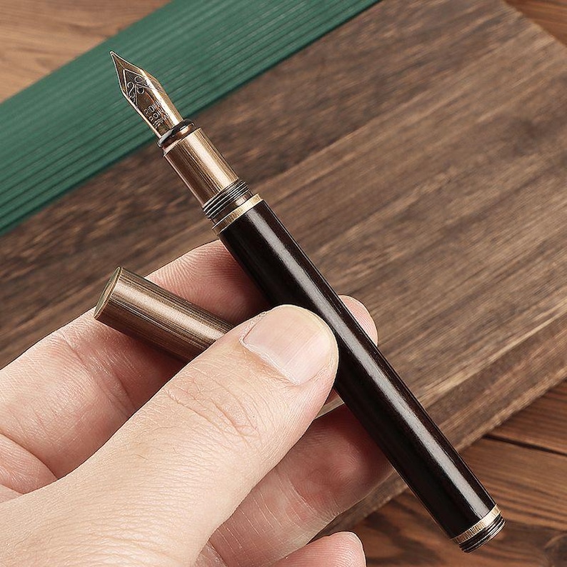 May include: A dark brown fountain pen with a gold-colored nib and accents. The pen's cap is detached and lies next to the pen. The pen is held against a wooden surface.