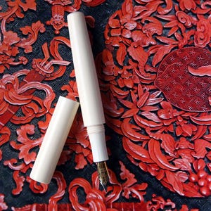 May include: A white fountain pen with a gold nib, resting on a red and black patterned surface. The pen is made of two sections that screw together.